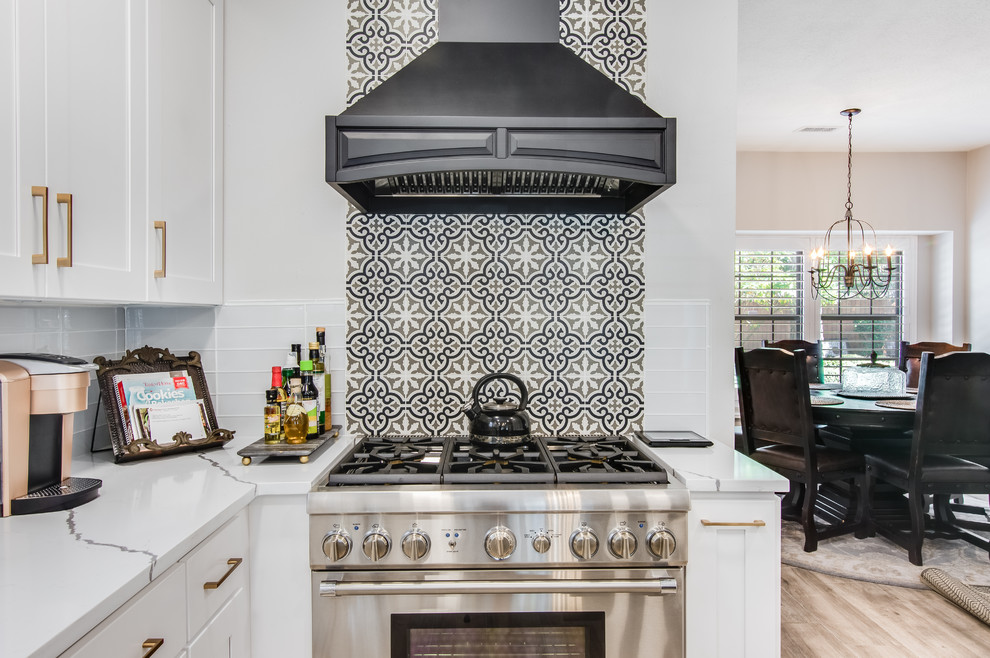Example of a large transitional l-shaped porcelain tile and gray floor kitchen pantry design in Houston with a farmhouse sink, shaker cabinets, white cabinets, quartzite countertops, white backsplash, glass tile backsplash, stainless steel appliances, an island and white countertops