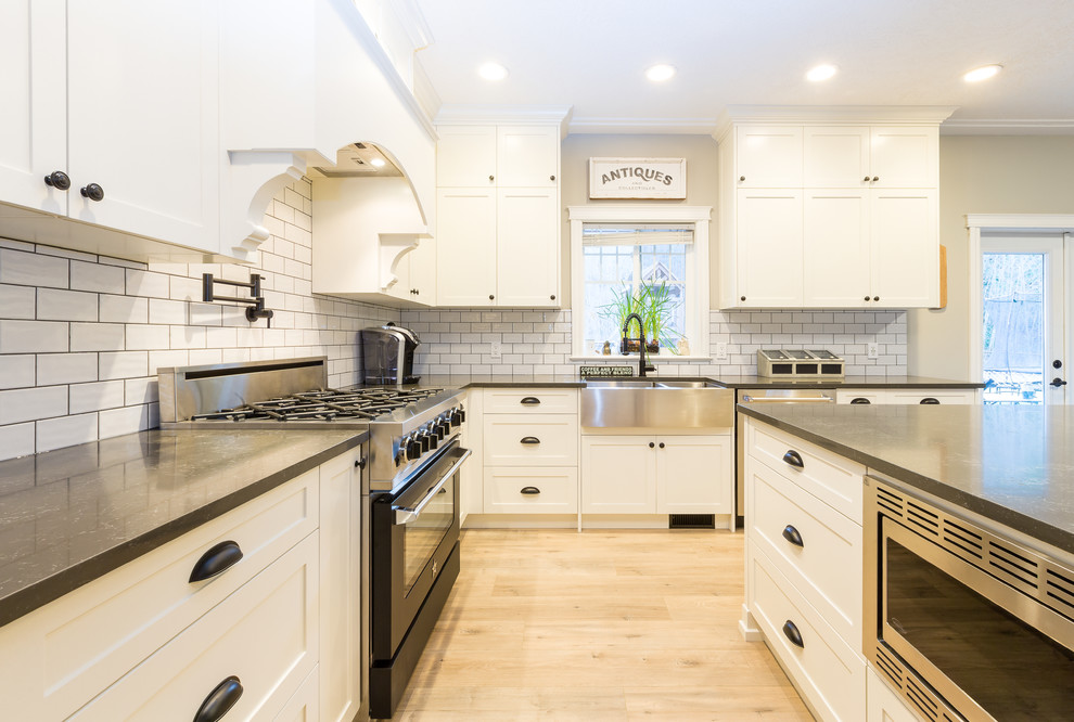 Photo of a large country l-shaped open plan kitchen in Other with a farmhouse sink, shaker cabinets, white cabinets, quartz benchtops, white splashback, subway tile splashback, stainless steel appliances, light hardwood floors, with island and black benchtop.