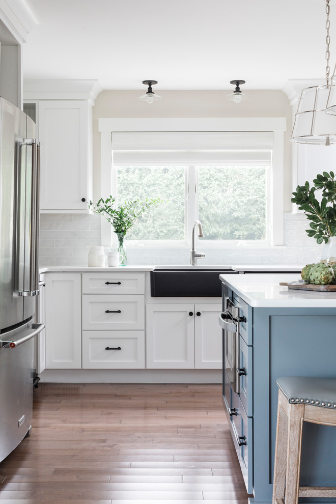 Photo of a large beach style l-shaped eat-in kitchen in Boston with a farmhouse sink, raised-panel cabinets, blue cabinets, quartz benchtops, white splashback, glass tile splashback, stainless steel appliances, medium hardwood floors, with island, grey floor and white benchtop.