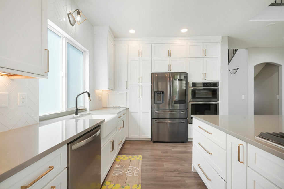 Large transitional l-shaped open concept kitchen photo in Denver with a farmhouse sink, shaker cabinets, white cabinets, quartz countertops, white backsplash, ceramic backsplash, stainless steel appliances, an island and gray countertops