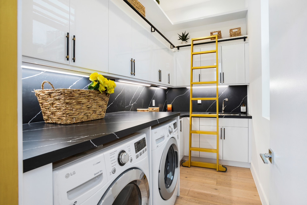 This is an example of a mid-sized contemporary l-shaped dedicated laundry room in Los Angeles with flat-panel cabinets, white cabinets, light hardwood floors, a side-by-side washer and dryer and black benchtop.