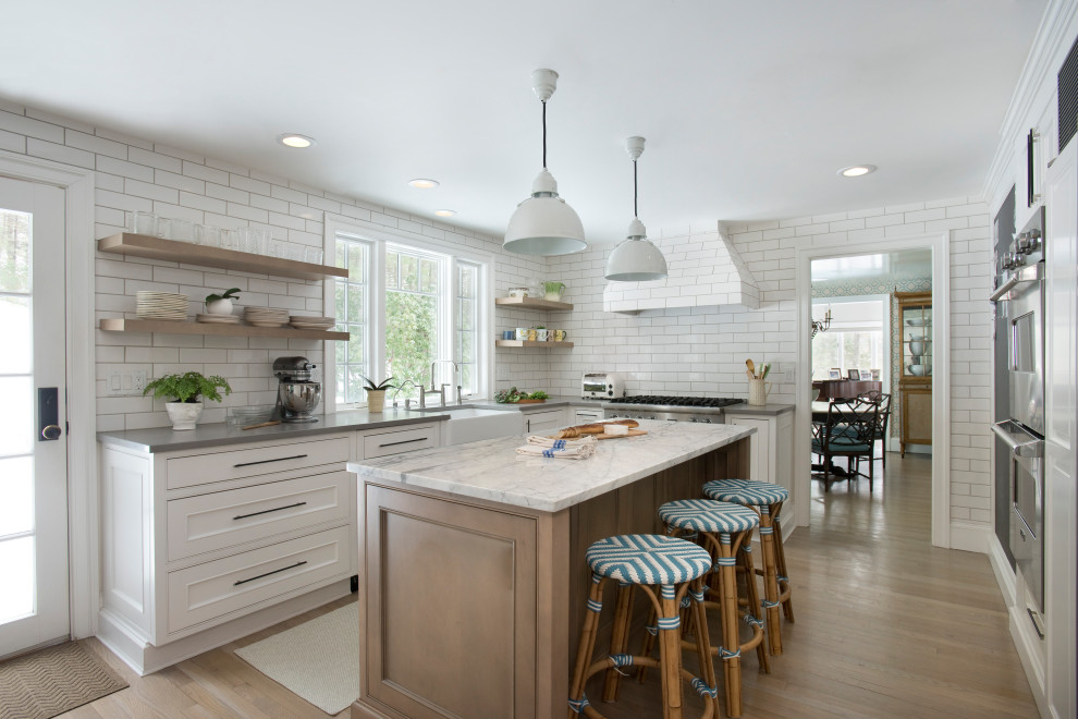 Example of a mid-sized transitional light wood floor and beige floor kitchen design in Boston with a farmhouse sink, shaker cabinets, quartz countertops, white backsplash, white appliances, an island, gray countertops, white cabinets and subway tile backsplash