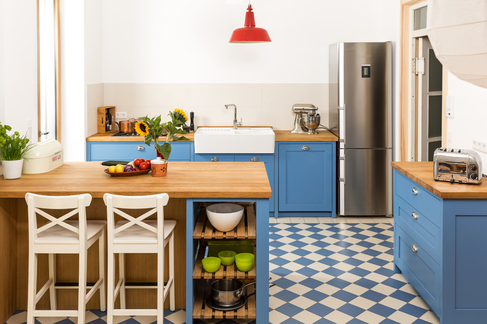 This is an example of a mid-sized country single-wall separate kitchen in Cologne with a farmhouse sink, shaker cabinets, blue cabinets, wood benchtops, grey splashback, ceramic splashback, a peninsula, stainless steel appliances and linoleum floors.