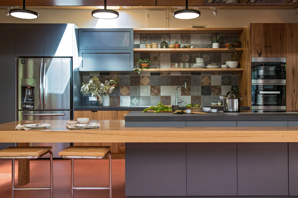 Inspiration for an expansive industrial galley open plan kitchen in Other with a double-bowl sink, flat-panel cabinets, medium wood cabinets, solid surface benchtops, multi-coloured splashback, ceramic splashback, stainless steel appliances, linoleum floors, with island, red floor and black benchtop.