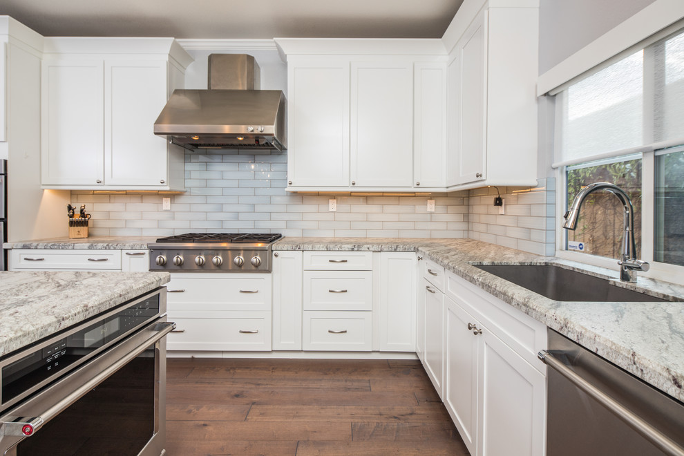 Photo of a large modern l-shaped kitchen in San Diego with an undermount sink, shaker cabinets, white cabinets, granite benchtops, grey splashback, glass tile splashback, stainless steel appliances, dark hardwood floors, with island and brown floor.