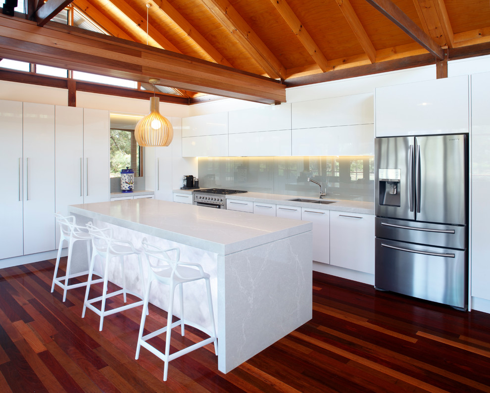 Inspiration for a tropical l-shaped kitchen in Newcastle - Maitland with a double-bowl sink, flat-panel cabinets, white cabinets, grey splashback, glass sheet splashback, stainless steel appliances, dark hardwood floors and with island.