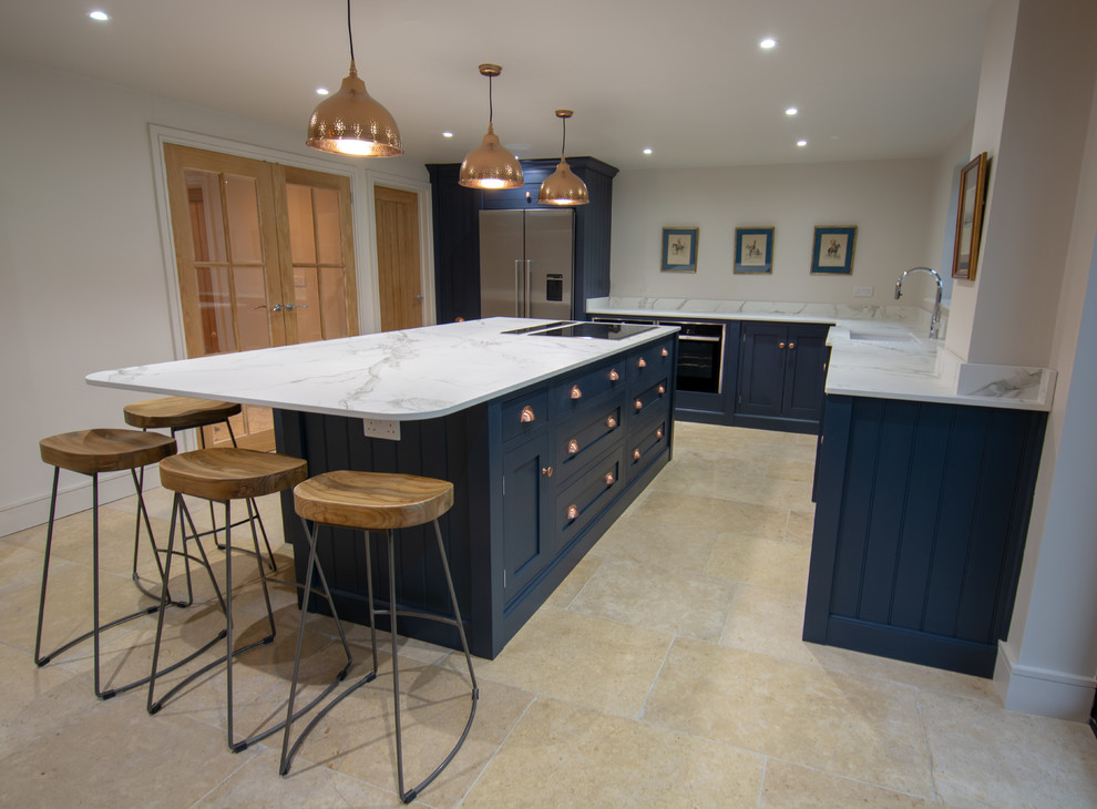 Photo of a mid-sized traditional l-shaped open plan kitchen in Sussex with an integrated sink, shaker cabinets, blue cabinets, solid surface benchtops, white splashback, stainless steel appliances, limestone floors, with island, beige floor and white benchtop.