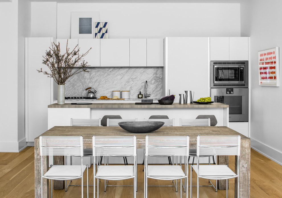 Inspiration for a mid-sized contemporary single-wall eat-in kitchen in New York with an undermount sink, flat-panel cabinets, white cabinets, white splashback, stone slab splashback, stainless steel appliances, light hardwood floors and with island.
