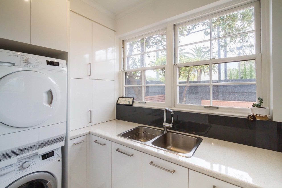 Photo of a small modern l-shaped utility room in Melbourne with a double-bowl sink, flat-panel cabinets, white cabinets, laminate benchtops, white walls, light hardwood floors and a stacked washer and dryer.