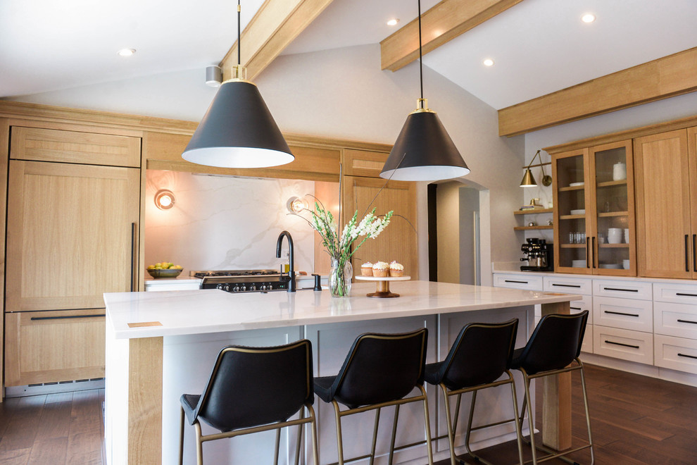 This is an example of a transitional l-shaped kitchen in New York with a farmhouse sink, shaker cabinets, light wood cabinets, white splashback, black appliances, dark hardwood floors, with island, brown floor and white benchtop.