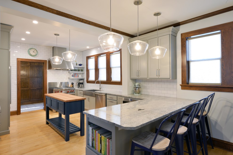 Example of a mid-sized transitional light wood floor and brown floor enclosed kitchen design with a farmhouse sink, shaker cabinets, gray cabinets, wood countertops, white backsplash, porcelain backsplash, stainless steel appliances, an island and brown countertops