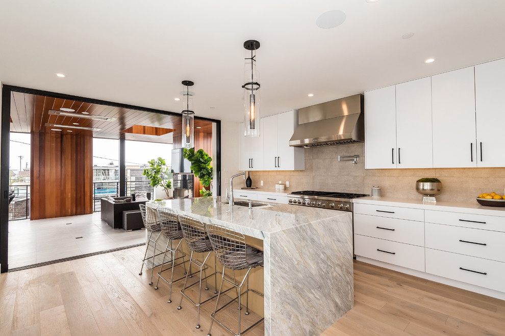 Photo of a contemporary galley kitchen in Los Angeles with an undermount sink, flat-panel cabinets, white cabinets, beige splashback, stainless steel appliances, light hardwood floors, with island, beige floor and white benchtop.