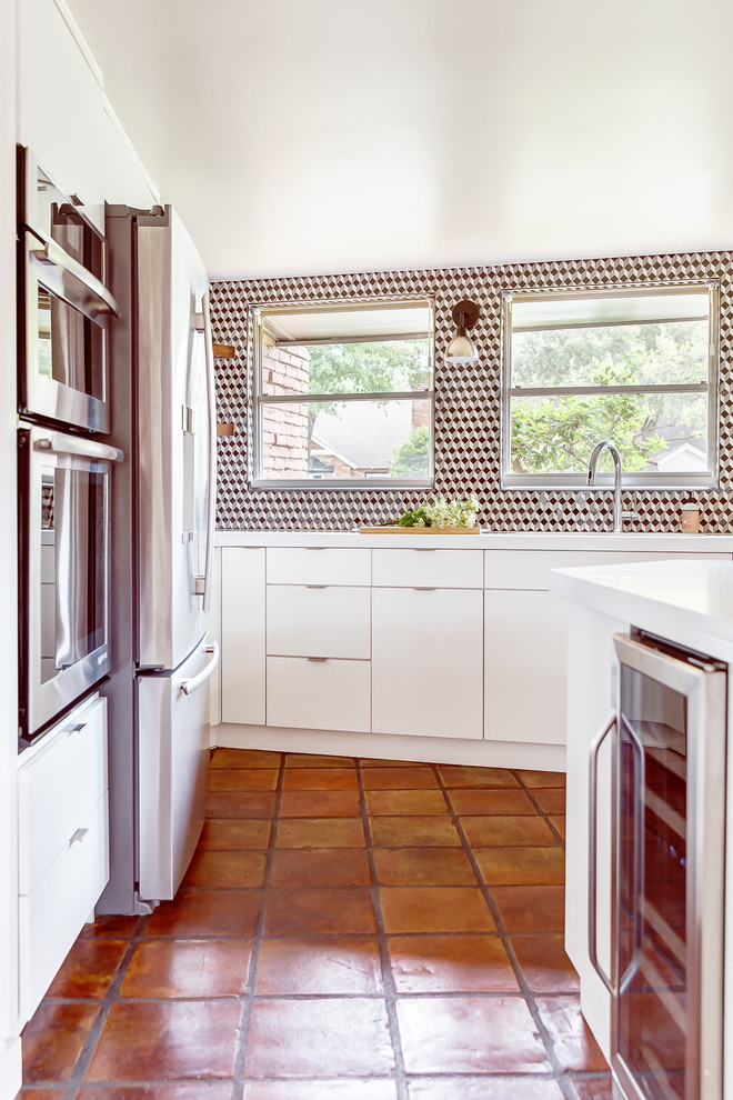 This is an example of a large midcentury galley kitchen in Houston with a double-bowl sink, flat-panel cabinets, white cabinets, solid surface benchtops, multi-coloured splashback, mosaic tile splashback, stainless steel appliances, terra-cotta floors, with island, brown floor and white benchtop.