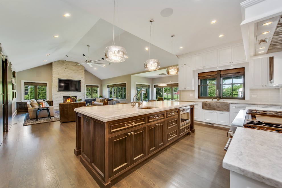 This is an example of a country u-shaped open plan kitchen in Chicago with a farmhouse sink, recessed-panel cabinets, white cabinets, marble benchtops, white splashback, porcelain splashback, stainless steel appliances, dark hardwood floors, with island and brown floor.