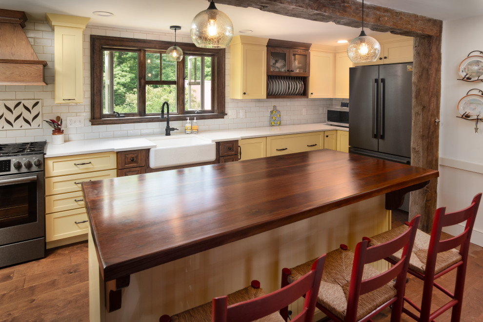 Photo of a large country l-shaped kitchen in Portland Maine with a farmhouse sink, shaker cabinets, yellow cabinets, quartz benchtops, white splashback, subway tile splashback, stainless steel appliances, dark hardwood floors, with island, brown floor and white benchtop.