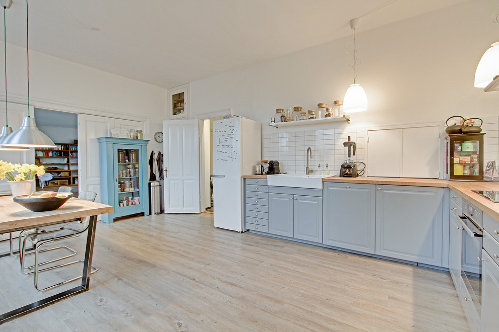 This is an example of a large scandinavian l-shaped eat-in kitchen in Aalborg with a farmhouse sink, raised-panel cabinets, grey cabinets, wood benchtops, white splashback, white appliances, light hardwood floors and no island.
