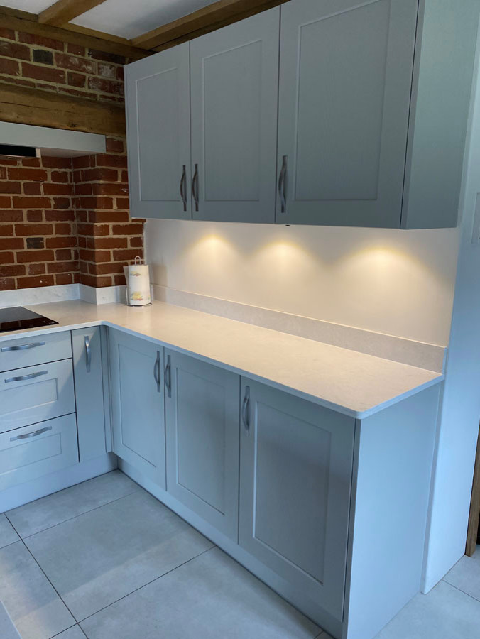 This is an example of a large country l-shaped open plan kitchen in Essex with an integrated sink, shaker cabinets, grey cabinets, quartz benchtops, white splashback, black appliances, ceramic floors, with island, white floor and white benchtop.