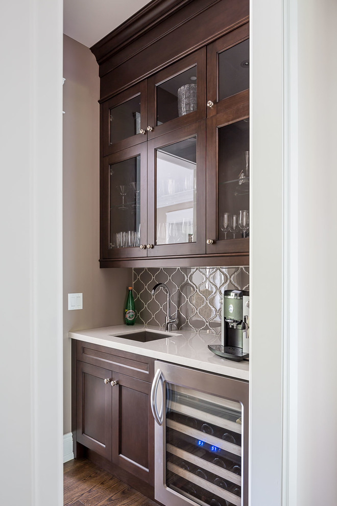 This is an example of a traditional wet bar in Toronto with an undermount sink, glass-front cabinets, dark wood cabinets, grey splashback and dark hardwood floors.