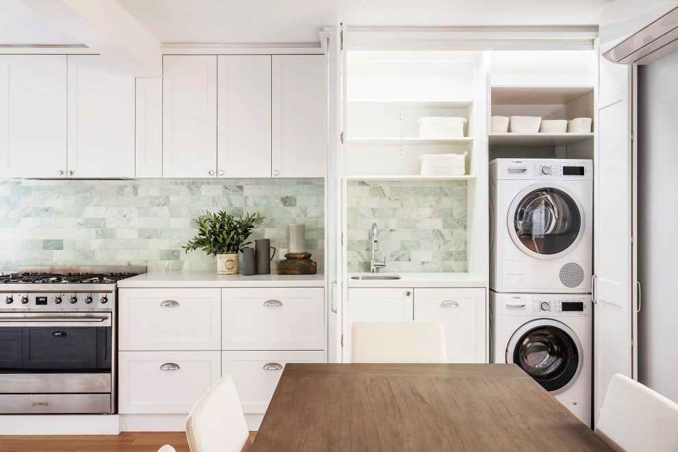 Design ideas for a small modern single-wall dedicated laundry room in Sydney with a single-bowl sink, shaker cabinets, white cabinets, marble benchtops, green splashback, subway tile splashback, white walls, medium hardwood floors, a stacked washer and dryer, brown floor and white benchtop.
