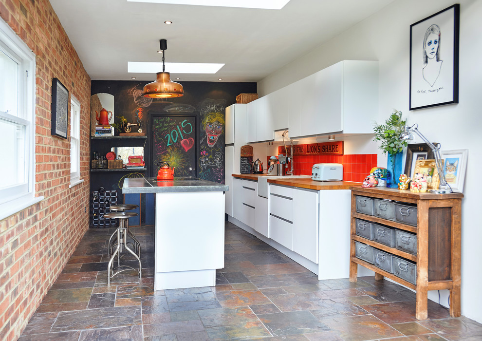 Mid-sized contemporary kitchen in London with a farmhouse sink, flat-panel cabinets, white cabinets, wood benchtops, orange splashback, ceramic splashback, stainless steel appliances, slate floors and with island.