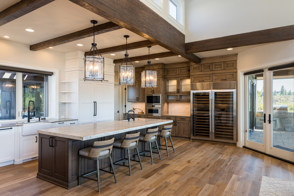 Large country l-shaped kitchen in Other with a farmhouse sink, shaker cabinets, white cabinets, quartzite benchtops, grey splashback, limestone splashback, stainless steel appliances, medium hardwood floors, with island, brown floor, white benchtop and exposed beam.