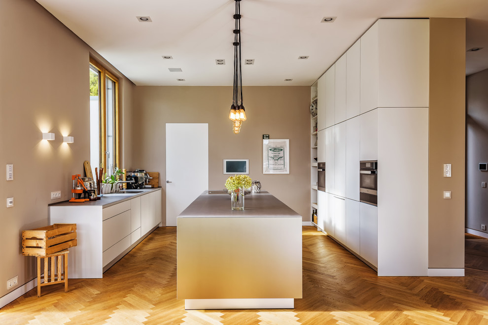 Photo of a large contemporary u-shaped open plan kitchen in Berlin with flat-panel cabinets, beige cabinets, stainless steel benchtops, medium hardwood floors, with island, brown floor, grey benchtop, an undermount sink and black appliances.