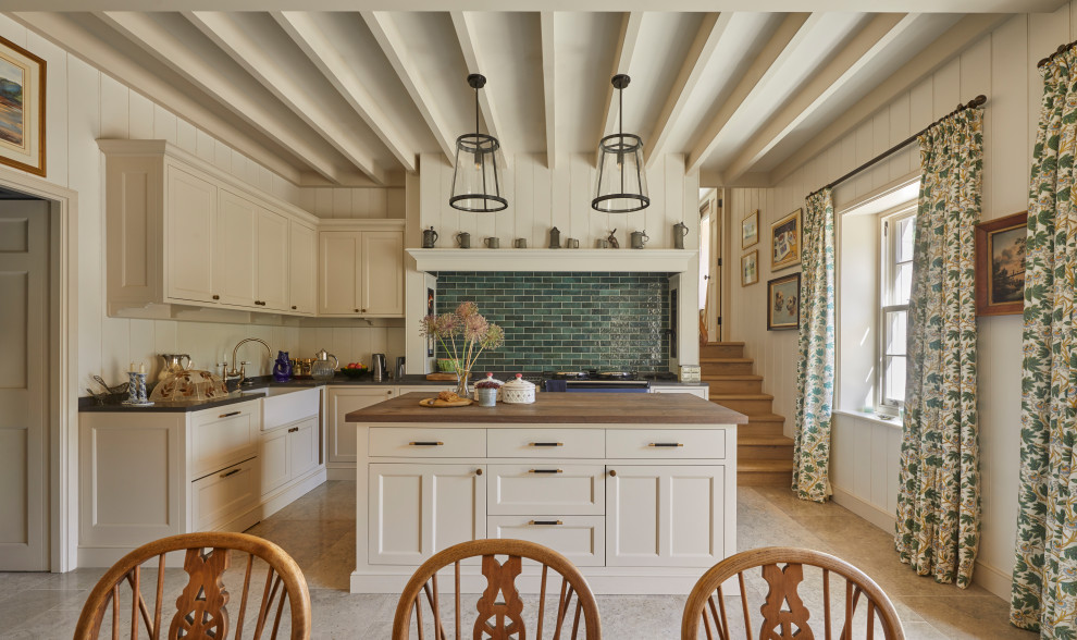 Example of a mid-sized country l-shaped limestone floor and brown floor open concept kitchen design in Wiltshire with a farmhouse sink, beige cabinets, wood countertops, green backsplash, ceramic backsplash, an island and brown countertops