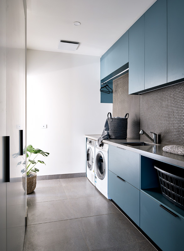 Photo of a large contemporary galley dedicated laundry room in Melbourne with an undermount sink, flat-panel cabinets, blue cabinets, solid surface benchtops, mosaic tile splashback, a side-by-side washer and dryer and grey benchtop.