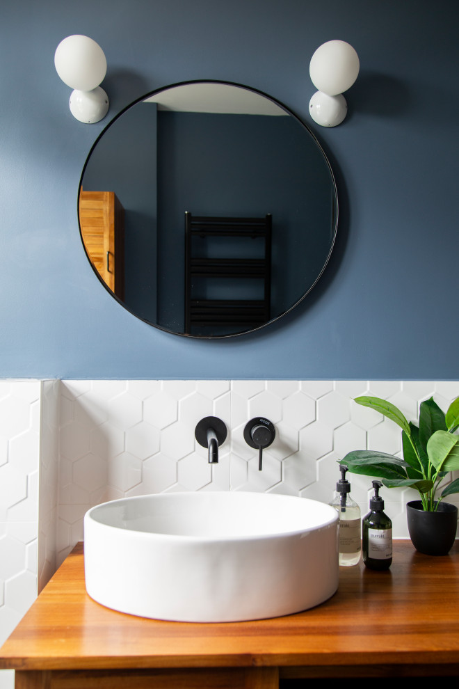 Photo of a small eclectic kids wet room bathroom in London with louvered cabinets, brown cabinets, a drop-in tub, a wall-mount toilet, white tile, porcelain tile, blue walls, porcelain floors, wood benchtops, black floor, brown benchtops, a single vanity, a freestanding vanity and wood.