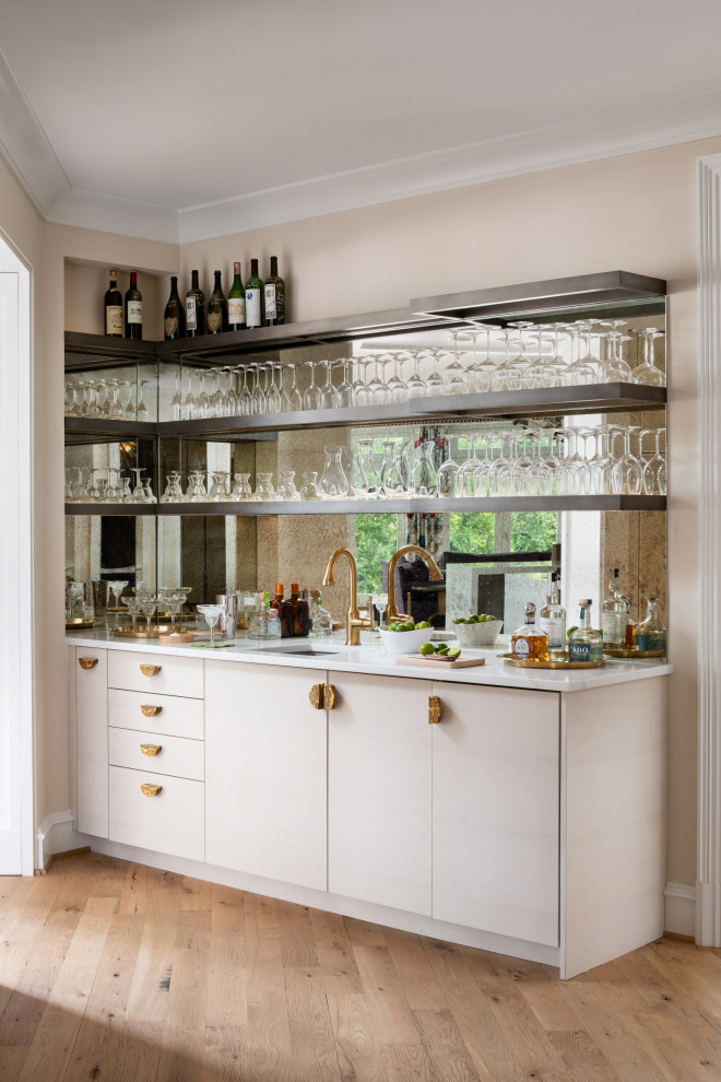 Photo of a transitional single-wall wet bar in DC Metro with an undermount sink, flat-panel cabinets, beige cabinets, mirror splashback, medium hardwood floors, brown floor and white benchtop.