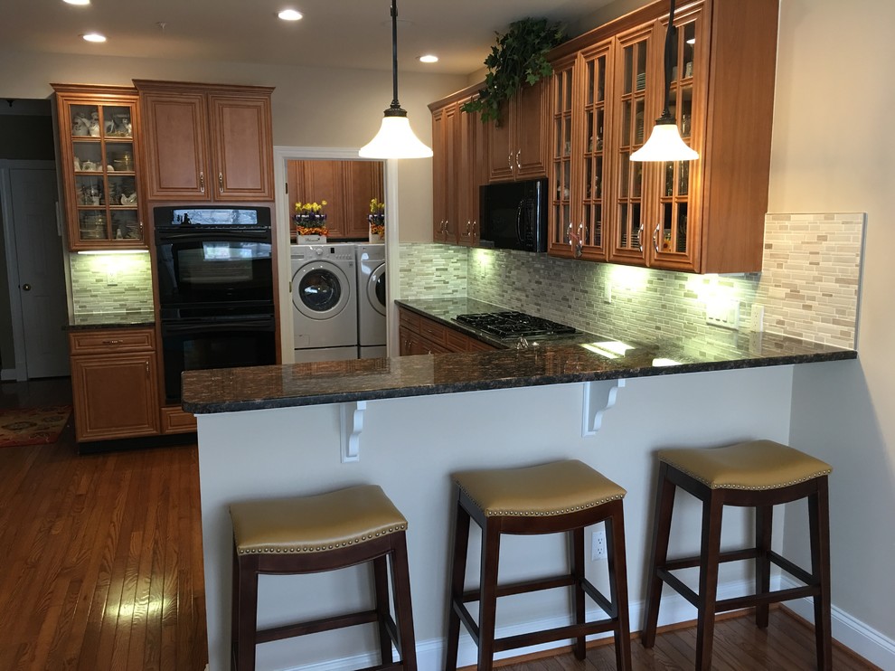 Mid-sized traditional u-shaped eat-in kitchen in Philadelphia with raised-panel cabinets, granite benchtops, grey splashback, matchstick tile splashback, black appliances, plywood floors, a peninsula, a double-bowl sink, medium wood cabinets and brown floor.