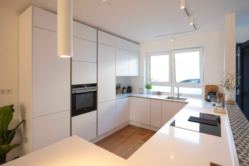 Inspiration for a mid-sized contemporary u-shaped open plan kitchen in Frankfurt with a drop-in sink, flat-panel cabinets, white cabinets, marble benchtops, white splashback, timber splashback, white appliances, medium hardwood floors, brown floor and white benchtop.