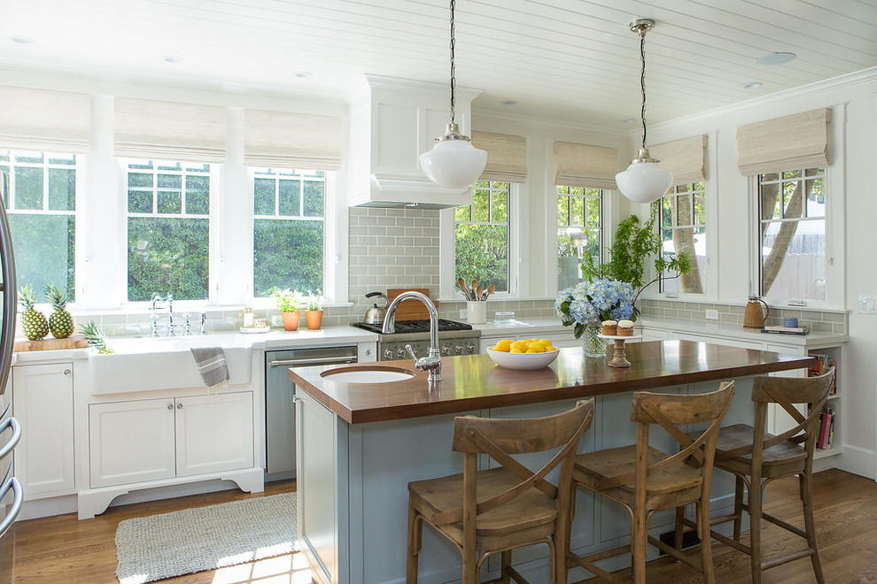 This is an example of a mid-sized beach style u-shaped eat-in kitchen in Santa Barbara with a farmhouse sink, shaker cabinets, white cabinets, wood benchtops, grey splashback, subway tile splashback, stainless steel appliances, medium hardwood floors, with island and brown floor.