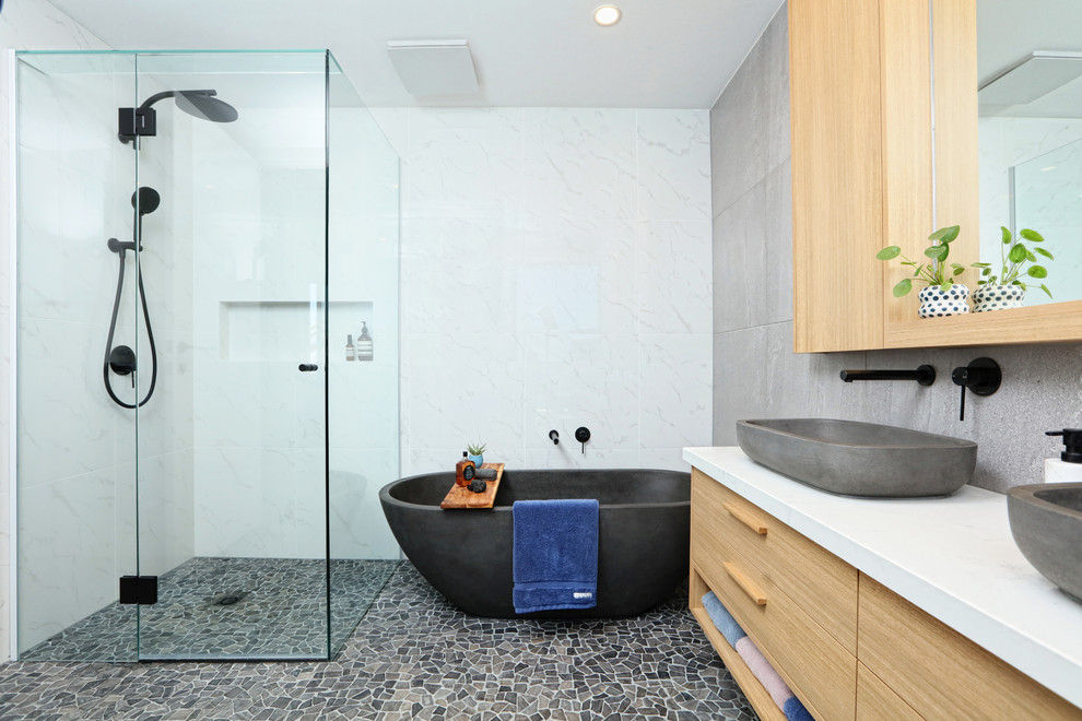 Photo of a mid-sized contemporary master bathroom in Melbourne with furniture-like cabinets, light wood cabinets, a freestanding tub, a one-piece toilet, porcelain tile, pebble tile floors, a vessel sink, engineered quartz benchtops and multi-coloured floor.