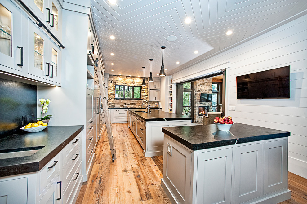 Large mountain style l-shaped medium tone wood floor and brown floor eat-in kitchen photo with an undermount sink, shaker cabinets, gray cabinets, soapstone countertops, black backsplash, stone slab backsplash, stainless steel appliances and two islands