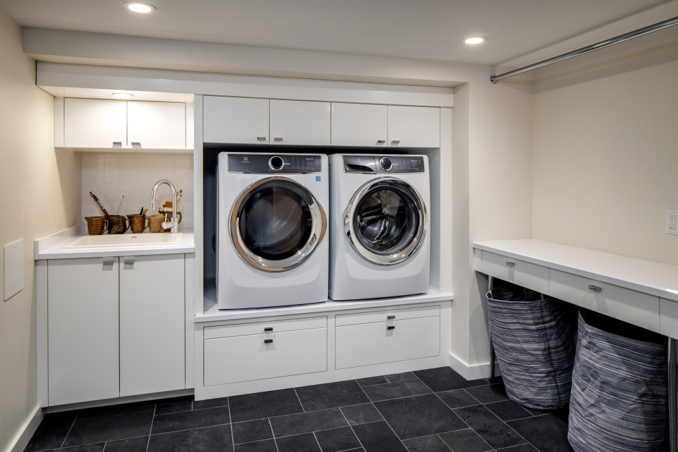 Idéer för en stor 60 tals vita linjär tvättstuga enbart för tvätt, med en nedsänkt diskho, släta luckor, vita skåp, bänkskiva i kvarts, vitt stänkskydd, stänkskydd i marmor, beige väggar, skiffergolv, en tvättmaskin och torktumlare bredvid varandra och svart golv
