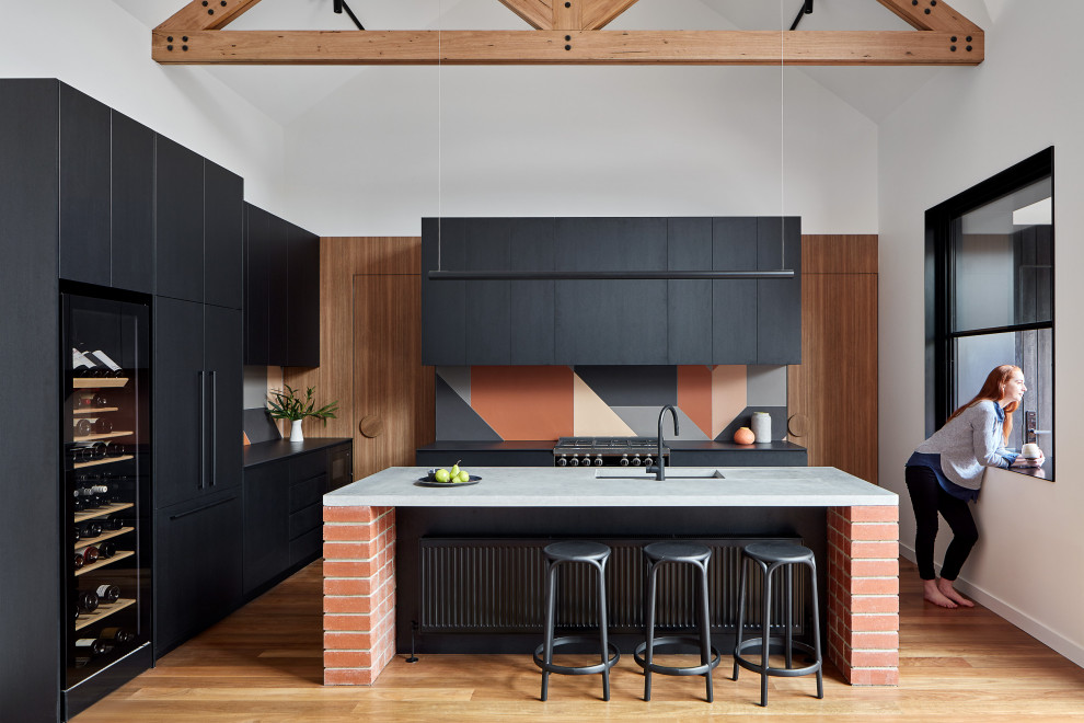 This is an example of a contemporary l-shaped kitchen in Melbourne with flat-panel cabinets, black cabinets, concrete benchtops, terra-cotta splashback, black appliances, medium hardwood floors, with island, black benchtop, an undermount sink, multi-coloured splashback, brown floor and vaulted.