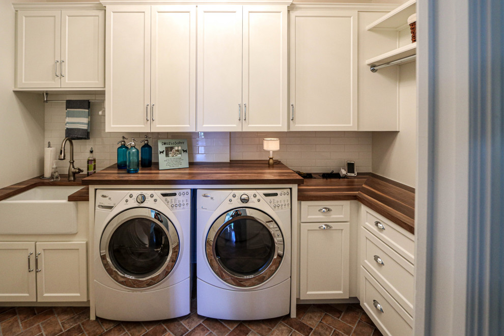 Photo of a small transitional l-shaped dedicated laundry room in Cleveland with a farmhouse sink, flat-panel cabinets, white cabinets, wood benchtops, white splashback, ceramic splashback, white walls, porcelain floors, a side-by-side washer and dryer, brown floor and brown benchtop.