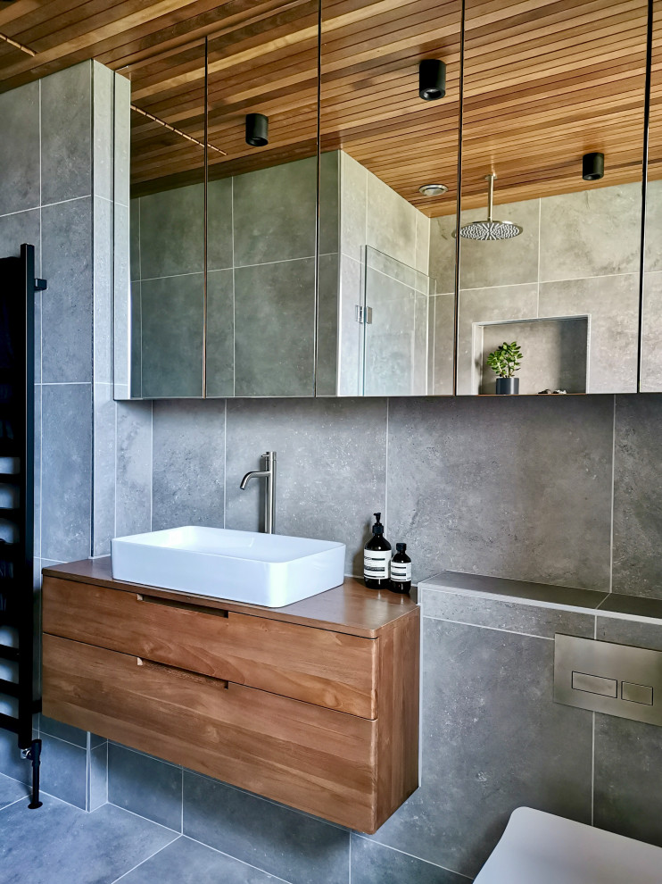 Photo of a small contemporary bathroom in Other with flat-panel cabinets, brown cabinets, a drop-in tub, a shower/bathtub combo, a wall-mount toilet, gray tile, porcelain tile, grey walls, porcelain floors, grey floor, a single vanity, a floating vanity and timber.