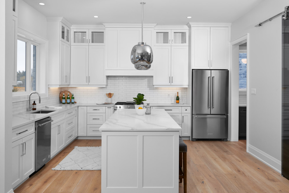 Example of a transitional l-shaped light wood floor and brown floor eat-in kitchen design in Toronto with an undermount sink, shaker cabinets, white cabinets, marble countertops, white backsplash, subway tile backsplash, stainless steel appliances, an island and white countertops