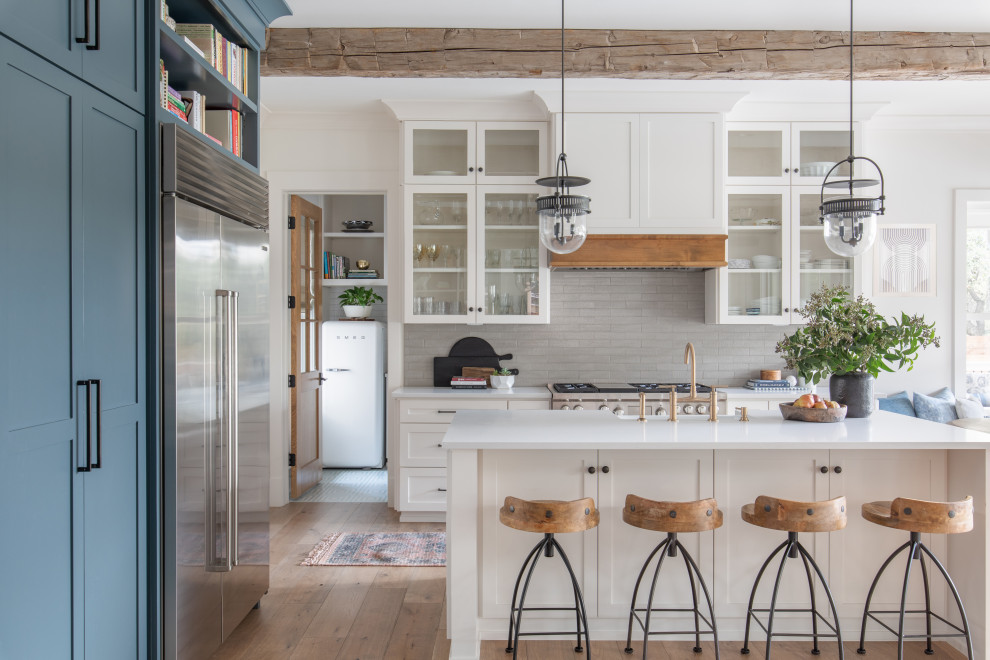 Example of a transitional u-shaped medium tone wood floor, brown floor and exposed beam open concept kitchen design in Austin with a farmhouse sink, shaker cabinets, white cabinets, quartz countertops, gray backsplash, stainless steel appliances, an island and white countertops