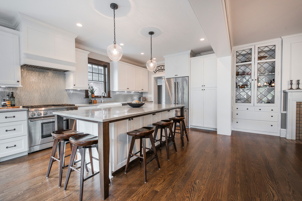 Photo of a large transitional l-shaped open plan kitchen in Columbus with shaker cabinets, white cabinets, quartzite benchtops, metallic splashback, glass tile splashback, stainless steel appliances, medium hardwood floors, with island, a farmhouse sink, brown floor and white benchtop.