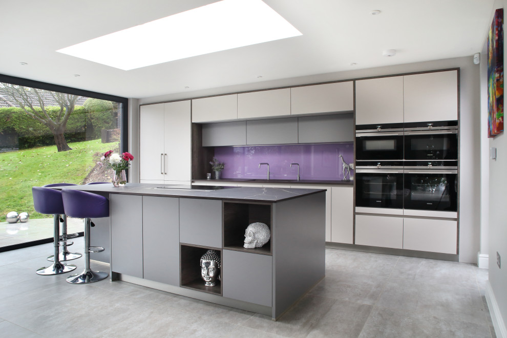 This is an example of a contemporary galley kitchen in Buckinghamshire with flat-panel cabinets, grey cabinets, glass sheet splashback, black appliances, with island, grey floor and black benchtop.