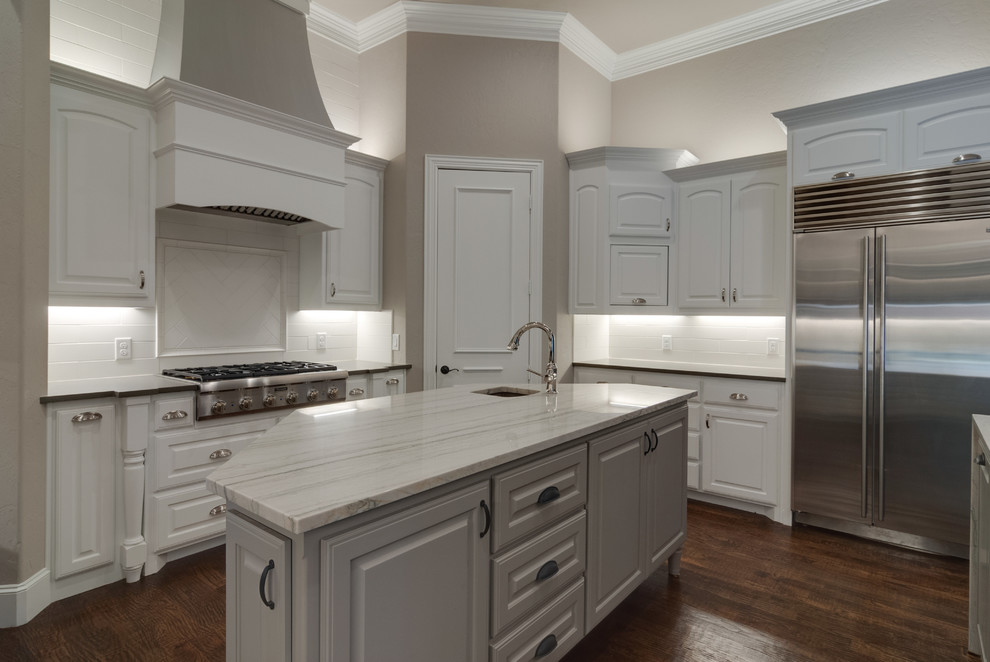 Mid-sized transitional u-shaped open plan kitchen in Dallas with an undermount sink, raised-panel cabinets, white cabinets, quartzite benchtops, white splashback, porcelain splashback, stainless steel appliances, medium hardwood floors, multiple islands, brown floor and white benchtop.