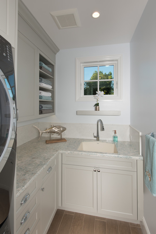 Small traditional l-shaped dedicated laundry room in San Francisco with an undermount sink, recessed-panel cabinets, grey cabinets, quartz benchtops, porcelain floors, a stacked washer and dryer, brown floor and grey walls.