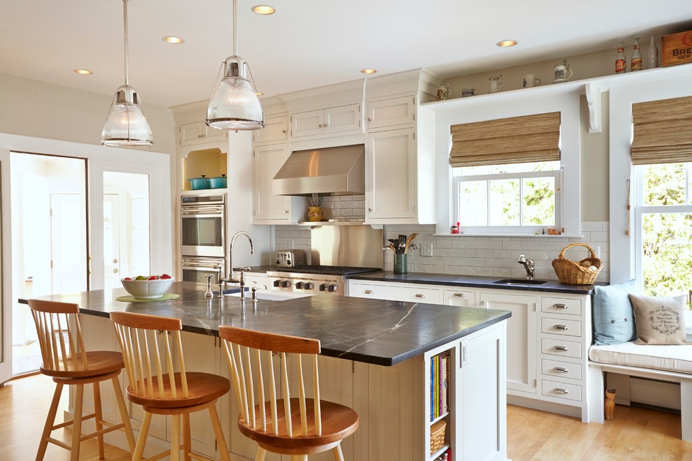 This is an example of a transitional galley kitchen in Boston with a farmhouse sink, shaker cabinets, white cabinets, soapstone benchtops, white splashback, subway tile splashback, stainless steel appliances, light hardwood floors and with island.