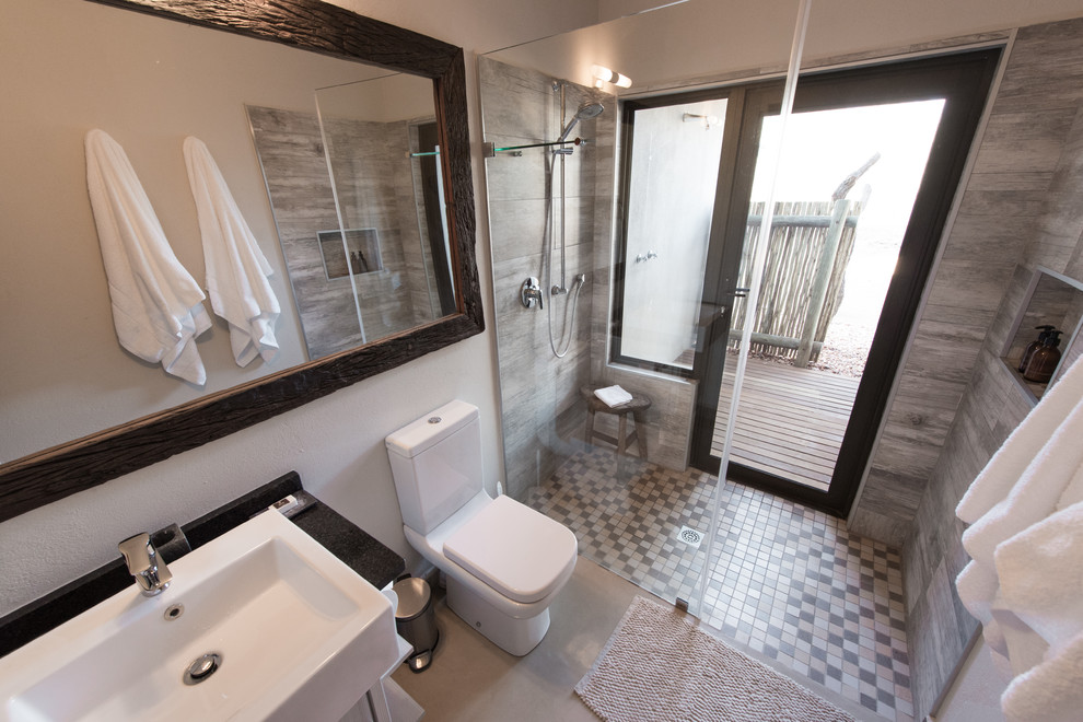 This is an example of a mid-sized country bathroom in Other with flat-panel cabinets, white cabinets, an open shower, an urinal, beige tile, ceramic tile, beige walls, concrete floors, a drop-in sink, granite benchtops, beige floor, a sliding shower screen and black benchtops.
