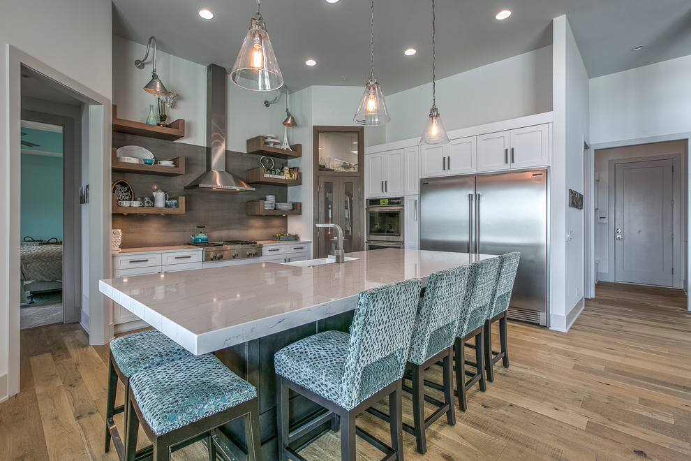 This is an example of a large beach style l-shaped kitchen in Omaha with a farmhouse sink, shaker cabinets, white cabinets, quartz benchtops, grey splashback, porcelain splashback, stainless steel appliances, with island, brown floor, white benchtop and medium hardwood floors.