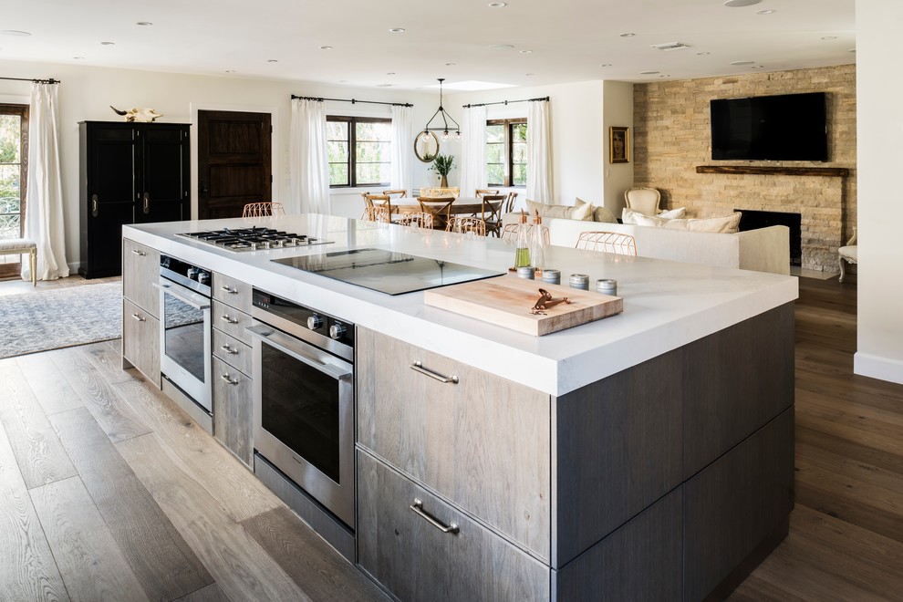Photo of a large contemporary l-shaped open plan kitchen in Los Angeles with an undermount sink, flat-panel cabinets, beige splashback, stone tile splashback, stainless steel appliances, light hardwood floors, with island, distressed cabinets and solid surface benchtops.