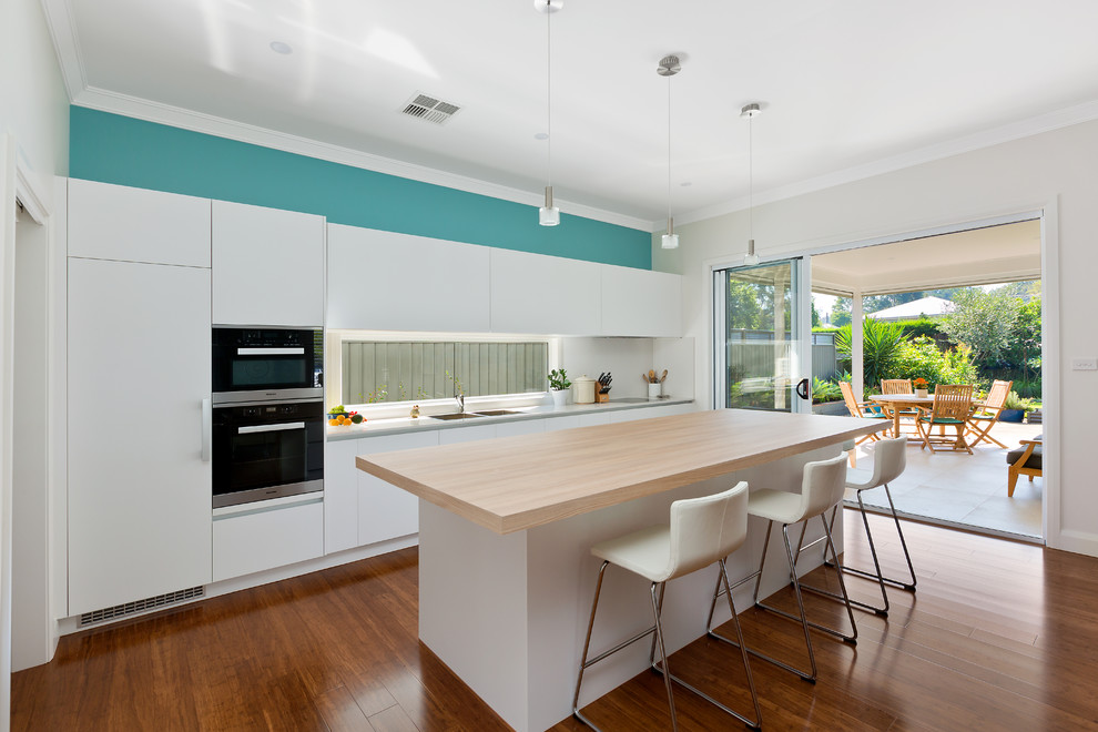 This is an example of a mid-sized beach style galley kitchen in Sydney with a drop-in sink, flat-panel cabinets, white cabinets, window splashback, black appliances, dark hardwood floors, with island, brown floor, wood benchtops and beige benchtop.